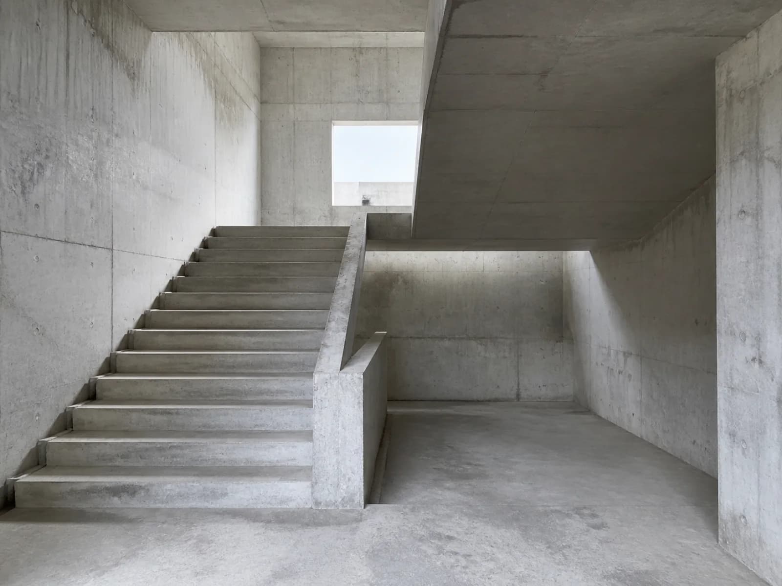 Empty concrete interior with a broad staircase, geometric landings, and natural light entering from above.